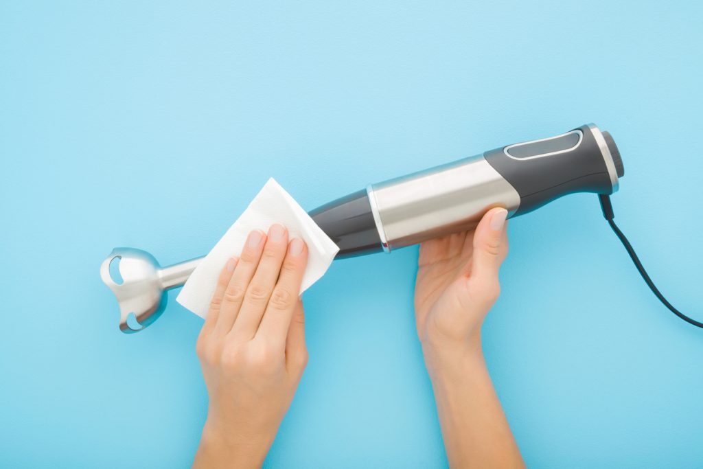 Young adult woman hands cleaning immersion blender with dry white paper napkin on light blue table background. Pastel color. Closeup. Home kitchen appliance purity. Point of view shot. Top down view.