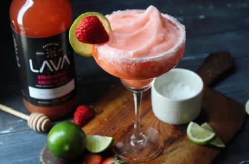 strawberry frozen margarita surrounded by lime, a cup of salt, and a bottle of strawberry cocktail mix